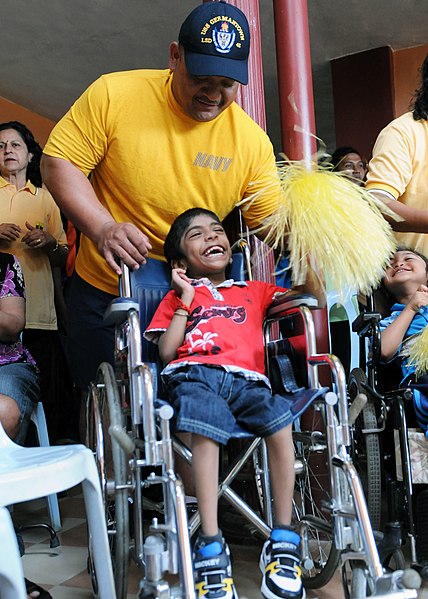 Un hombre con camisa amarilla se para detrás de un joven sonriente en silla de ruedas.