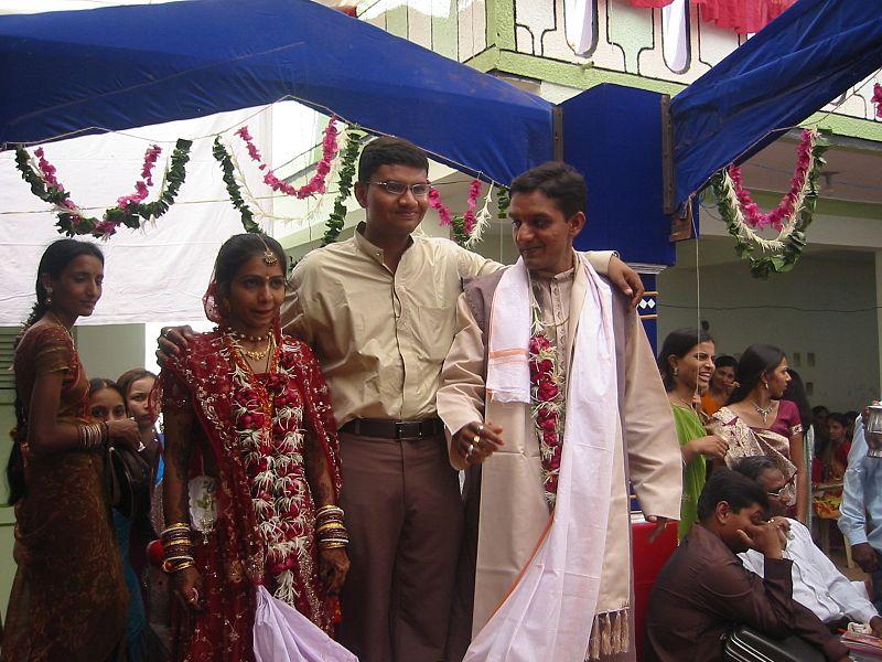 An indian wedding. The bride and groom are standing near each other and a long, white sash from the woman's gown is wrapped around the man's shoulders.