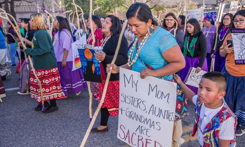 woman in protest holding sign that reads 'My mom, sisters, aunties + grandmas are sacred'