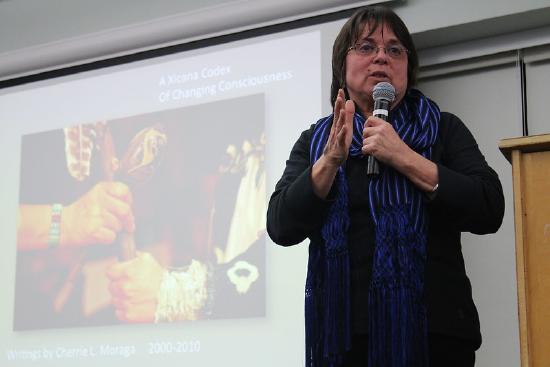 Cherrie Moraga wearing glasses and a scarf with a mic in her hand, speaking to an audience