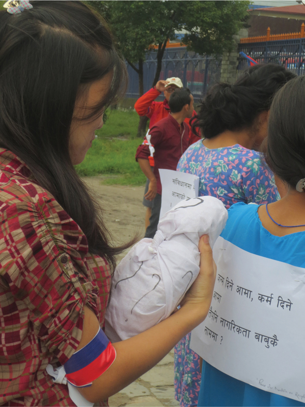 Figure 4.2. A young woman carries an effigy of a baby covered with question marks. The sign pinned to the back of the protester in front of her reads “Birth and karma are given by mothers. Why does citizenship have to be in the father’s name?” Tundikhel, Kathmandu, September 20, 2014. Dannah Dennis; CC BY.