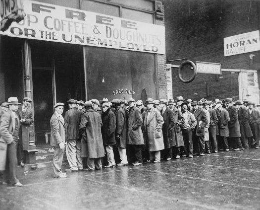 照片显示了一排穿着长外套和帽子的人在建筑物外排队，上面写着 “为失业者提供免费杯咖啡和甜甜圈” 的标语。