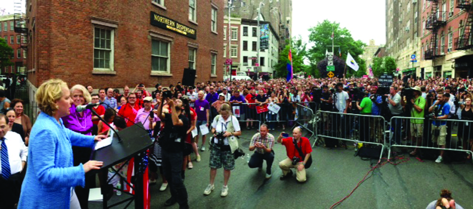 L'image montre deux personnes sur un podium devant une foule nombreuse dans une rue de la ville. Une personne s'adresse à la foule, tandis que l'autre se tient à côté du podium.
