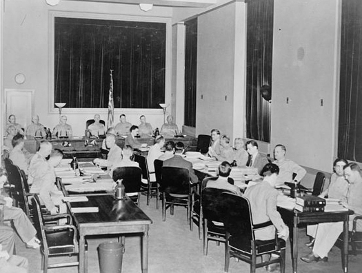 Uma foto de um grupo de pessoas em uma comissão militar, sentadas em cadeiras ao redor de várias mesas dispostas em forma de U.