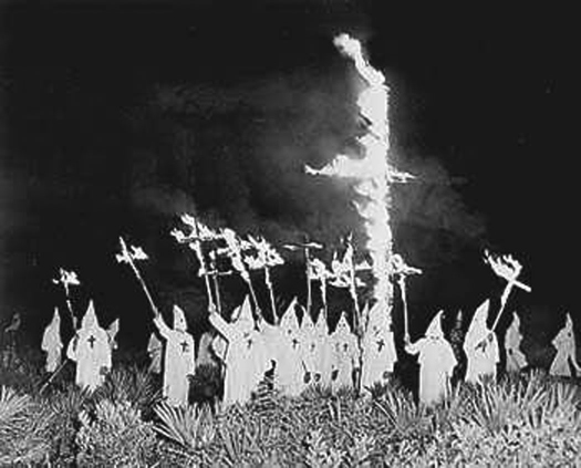 Photo d'un groupe de personnes portant des robes et des chapeaux pointus, entourant une grande croix en feu dans le sol. Plusieurs personnes brandissent des croix en feu en altitude.