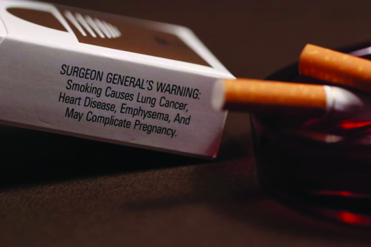 Uma foto de uma caixa de cigarros e dois cigarros. Os cigarros estão em um cinzeiro. O texto na caixa de cigarros diz “Aviso do cirurgião geral: fumar causa câncer de pulmão, doenças cardíacas, enfisema e pode complicar a gravidez”.