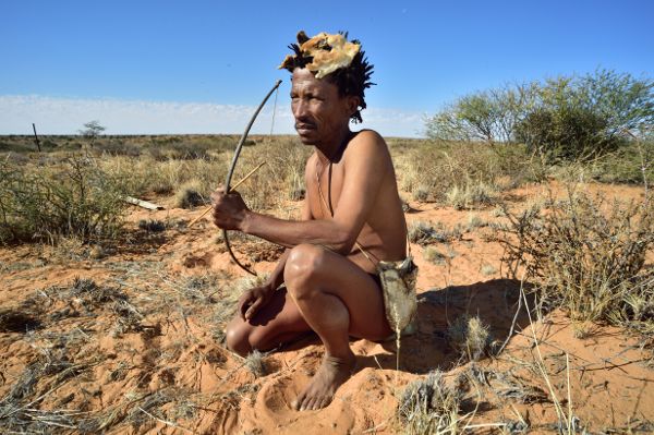 Arri Raats, Kalahari Khomani San Bushman, Boesmansrus camp, Northern Cape, South Africa is shown squatting in a dry, red earthed land, holding a small bow and arrow and wearing a animal skin on his head.