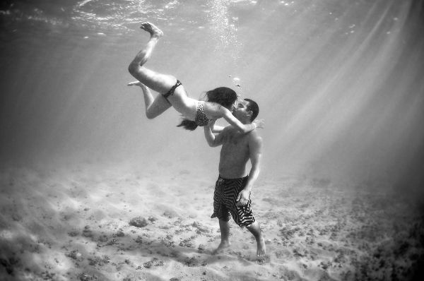 A guy is standing underwater and a girl is swimming down and is kissing him.