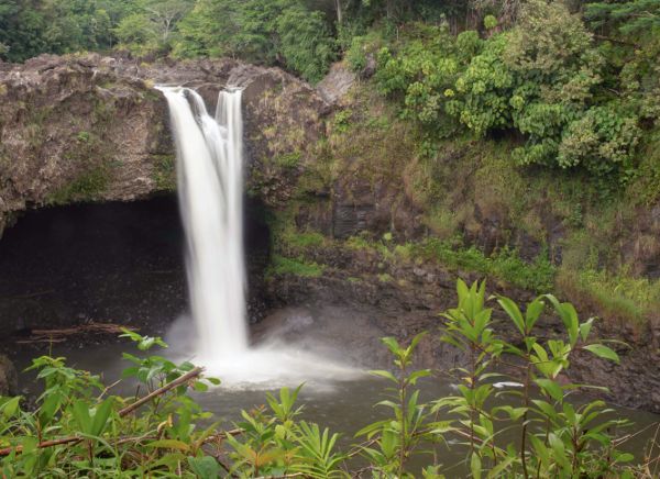 A waterfall in Hawaii splashes down into a pool in front of a deep cave.