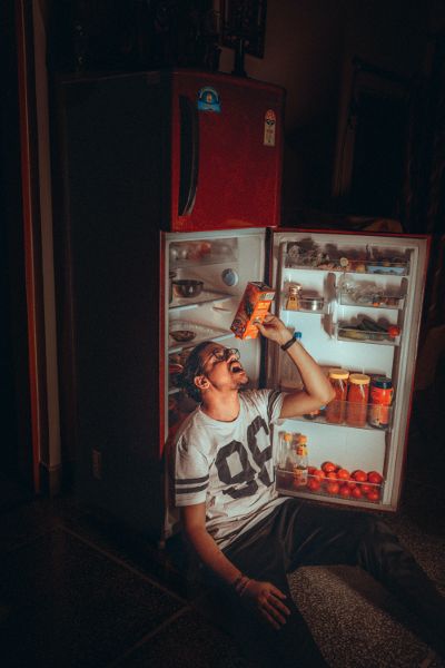 Un hombre está sentado frente a un refrigerador abierto por la noche mientras le vierte jugo en la boca.
