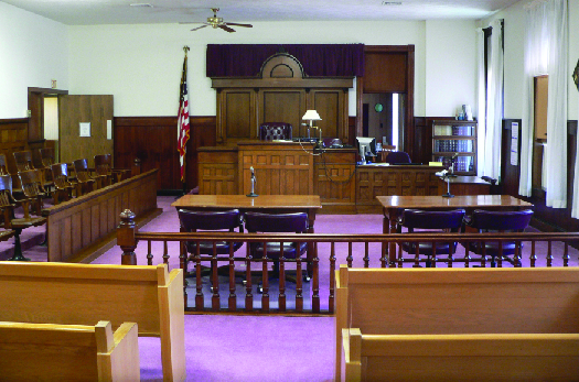 一张典型法庭的照片，里面没有人。 前景是供与会者使用的长椅，然后是辩方和检方的两张桌子，背景是法官的看台。 法官看台的左边是一排陪审团的椅子，法官席的右边是证人席。