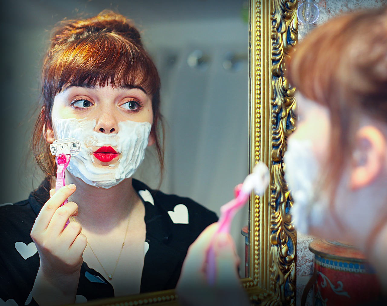 White-appearing person with red hair looking in the mirror. The person is shaving her face with a pink razor.