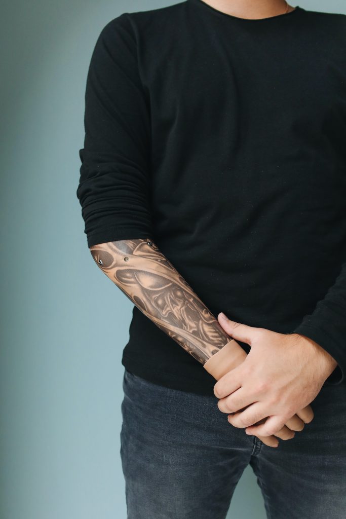 A person is standing with their hands clasped in front of them with a black sweater pulled down over where their prosthetic arm attaches at their elbow. The prosthetic is adorned with tattoos and matches the person's skin tone.