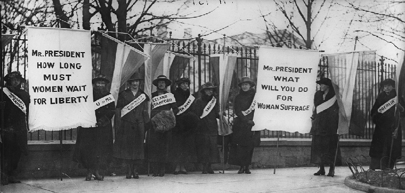 几个人站在栅栏前的照片。 有些人拿着横幅。 标语上写着 “总统先生，妇女要等多久才能获得自由” 和 “总统先生，你将为妇女受苦做些什么”。