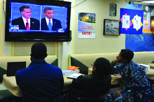 Uma imagem de três pessoas assistindo televisão. Na tela da televisão estão Mitt Romney e Barack Obama.