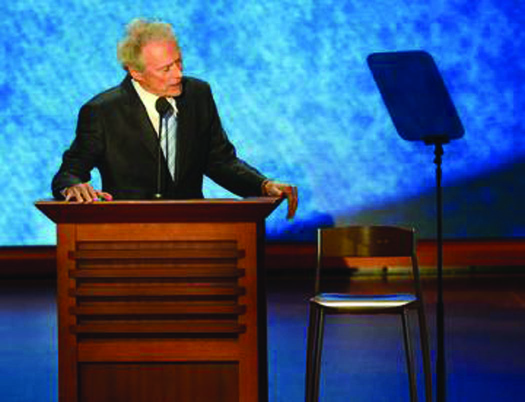 克林特·伊斯特伍德站在讲台后面的照片。 右边他旁边是一把空椅子。