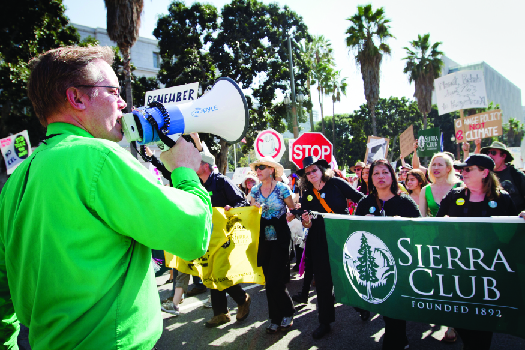 Image d'une personne parlant à travers un mégaphone sur la gauche et d'une foule de personnes marchant dans une rue sur la droite. Plusieurs marcheurs brandissent une grande banderole sur laquelle on peut lire « Sierra Club ».