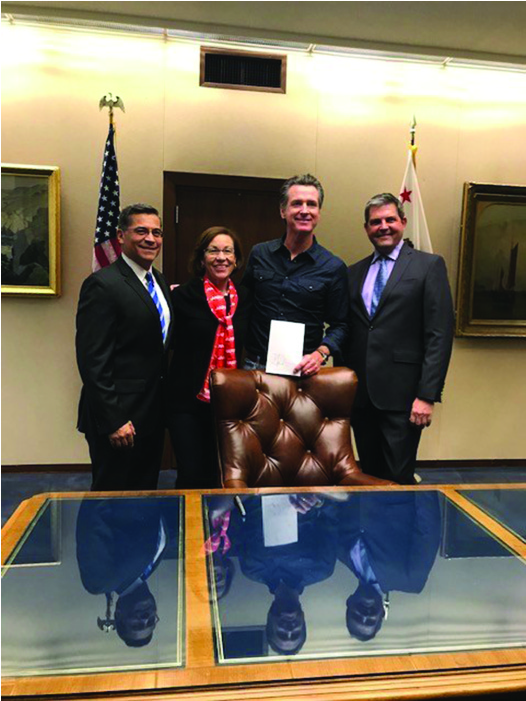 Four people stand behind a desk: (Left to Right) Attorney General Becerra, AARP Advocacy Manager Blanca Castro, Governor Newsom, and Assemblymember Jim Wood. Newsom holds a document.