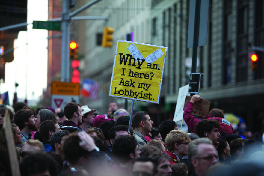Image d'une foule de personnes, dont l'une tient une pancarte sur laquelle on peut lire « Pourquoi suis-je ici ? Demandez à mon lobbyiste ».