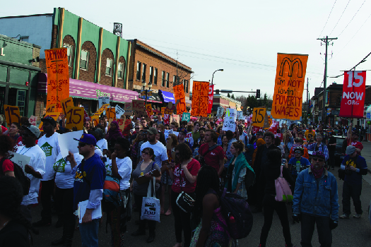 Image d'un groupe de personnes marchant dans une rue, dont l'une tient une pancarte sur laquelle on peut lire « Je n'aime pas les salaires de pauvreté ».