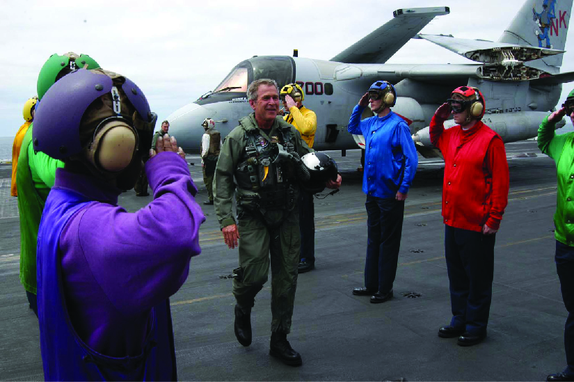 Une photo de George W. Bush dans une combinaison de vol sortant d'un avion pour monter sur un porte-avions. Le personnel se tient debout de chaque côté et le salue.