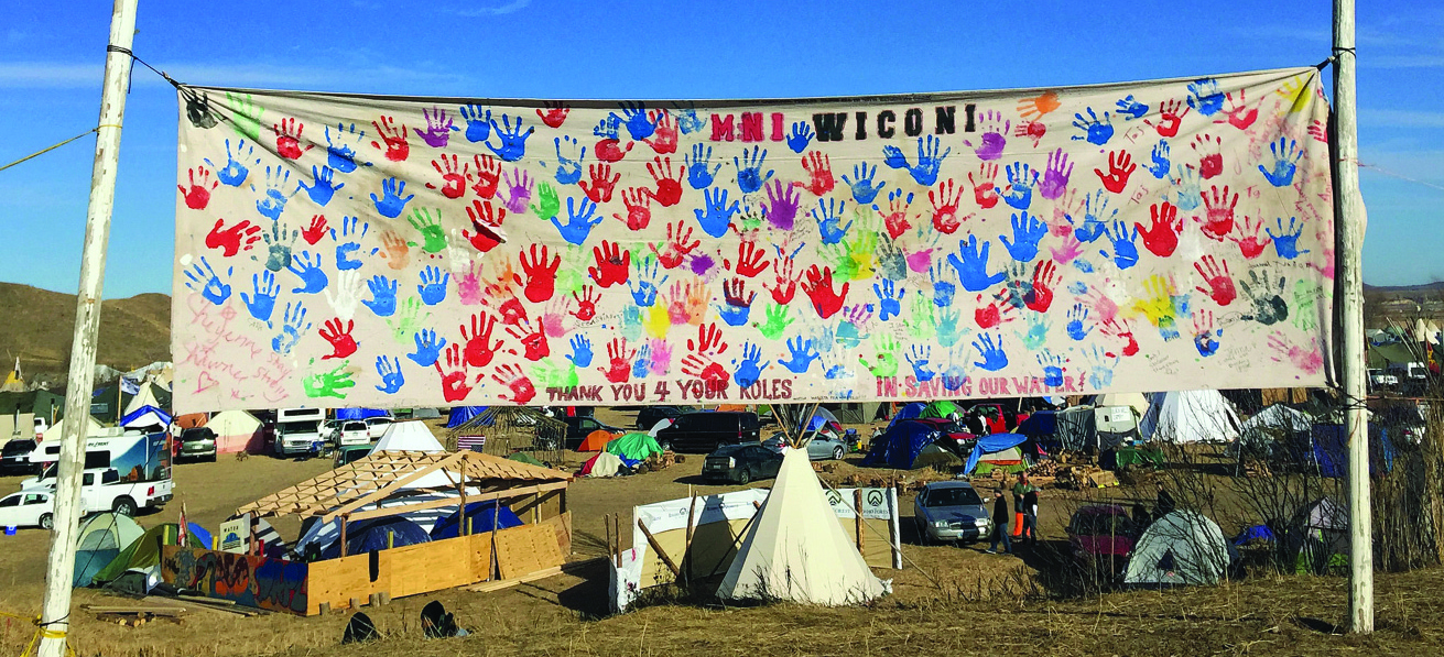 Um acampamento mostra vários veículos, tendas e uma tenda. Um grande banner coberto com impressões de mãos coloridas diz “MNI WICONI” e “Thank You 4 Your Roles in Saving Our Water”.