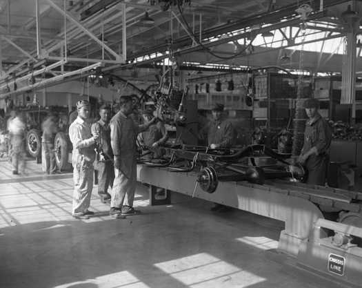Uma imagem de algumas pessoas paradas em uma fábrica automotiva ao lado de algumas máquinas.