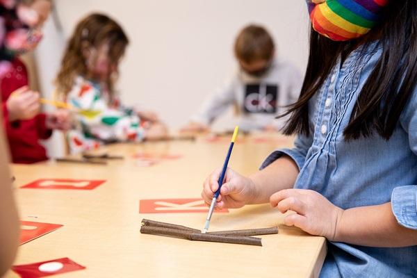 Los niños se sientan en una mesa con plantillas de letras y pegan letras. Rastrean las letras de palo con un pincel.