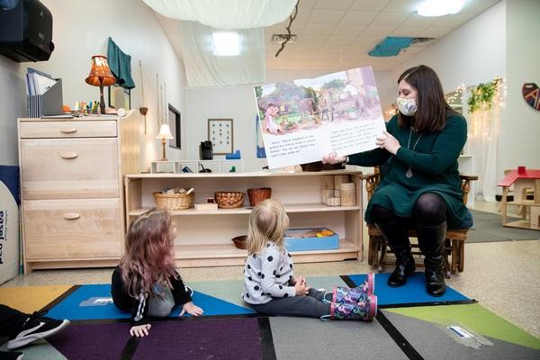Un maestro se sienta en una silla sosteniendo un libro grande con las fotos y la impresión apuntando hacia los dos niños sentados en la alfombra. Los dos niños miran el libro.