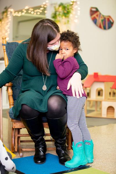 A teacher sits and comforts an upset child who is standing next to her.