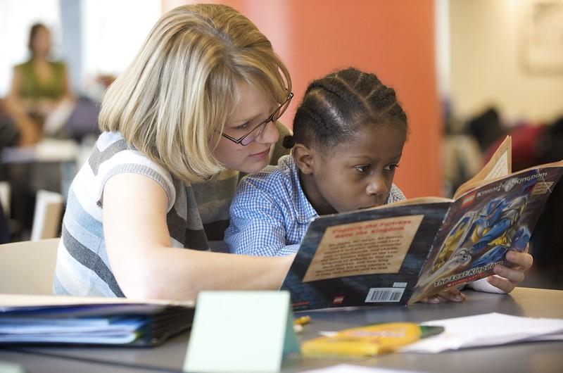 Un maestro se sienta detrás de un niño y señala las características del texto mientras el niño lee un libro en una mesa.