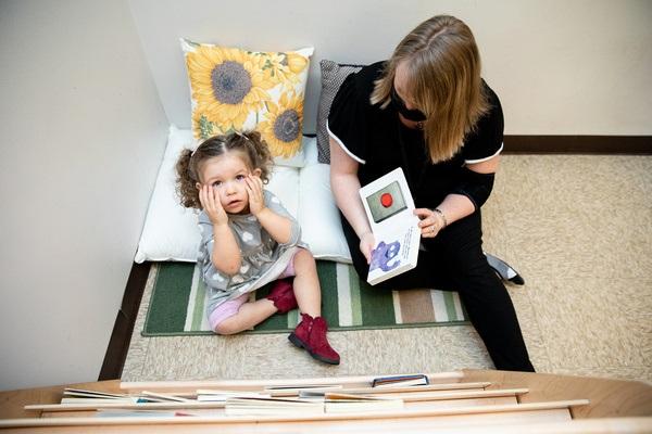 Un maestro se sienta en una almohada en el piso y muestra a un niño sentado en un tapete cuadros en un libro de cartón.