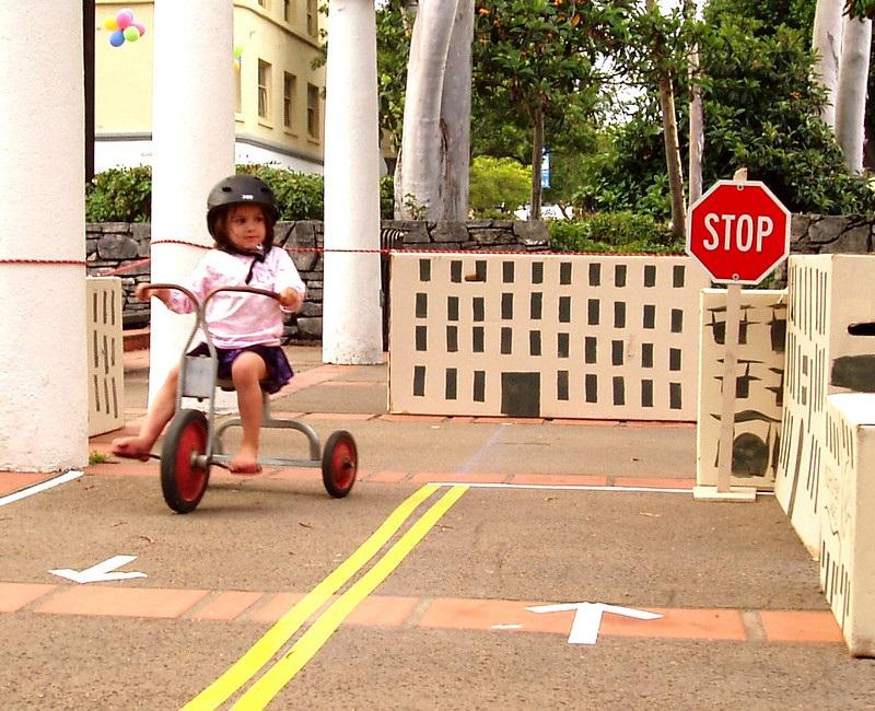 Un niño monta un triciclo por concreto que tiene flechas blancas que muestran en qué dirección andar a cada lado de una doble línea amarilla. Una señal de alto se encuentra al final de un lado de la carretera simulada.