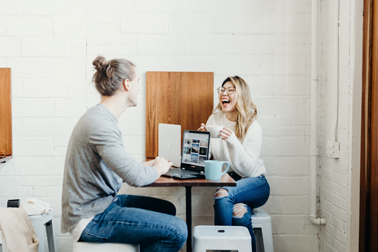 Two people at a table drinking coffee and laughing while working from laptops. 