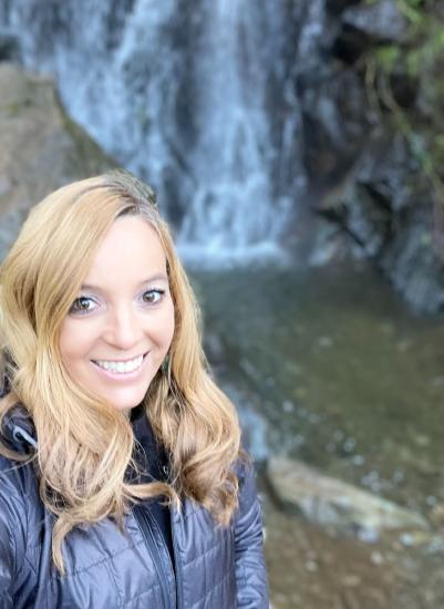 Elizabeth or Liz loves to hike and is posing in front of a waterfall in a blue jacket