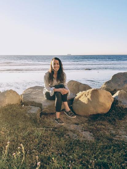 Alex is sitting on rocks with the ocean behind her. She has long brown hair and is in all blue