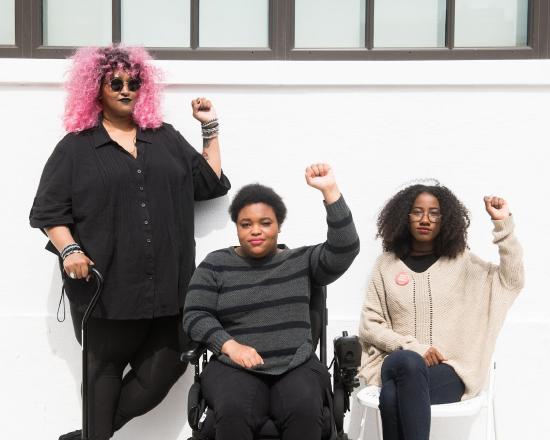 A non-binary person holding a cane, a non-binary person in a wheelchair, and a femme on a folding chair raising their fists