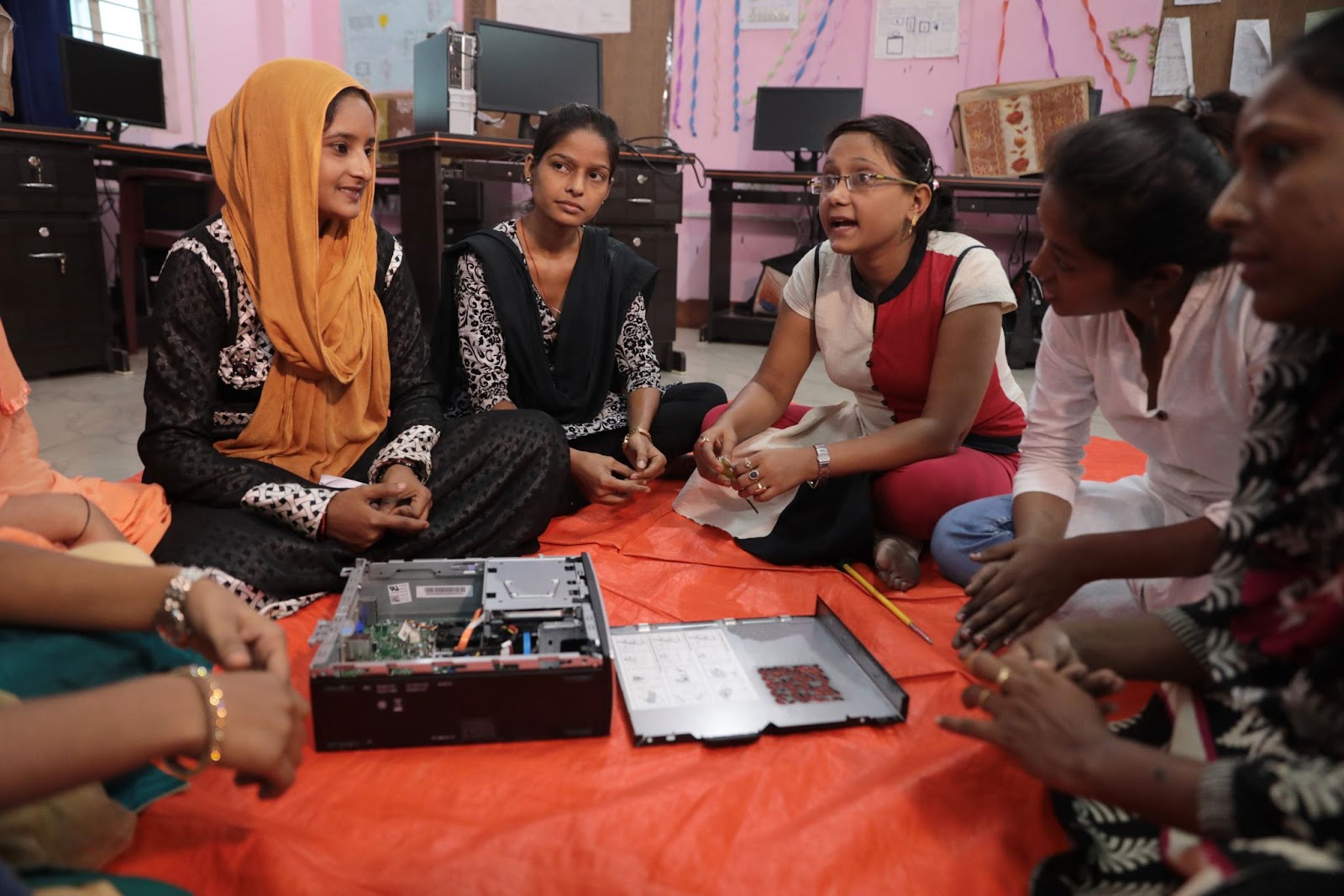 A group of women sit in a circle discussing computer hardware equipment. A few wear traditional Indian clothing.