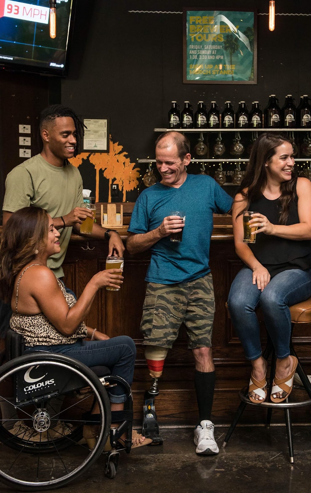 A group of people at a bar drinking and talking; one man has a prosthetic leg, and one woman sits in a wheelchair.