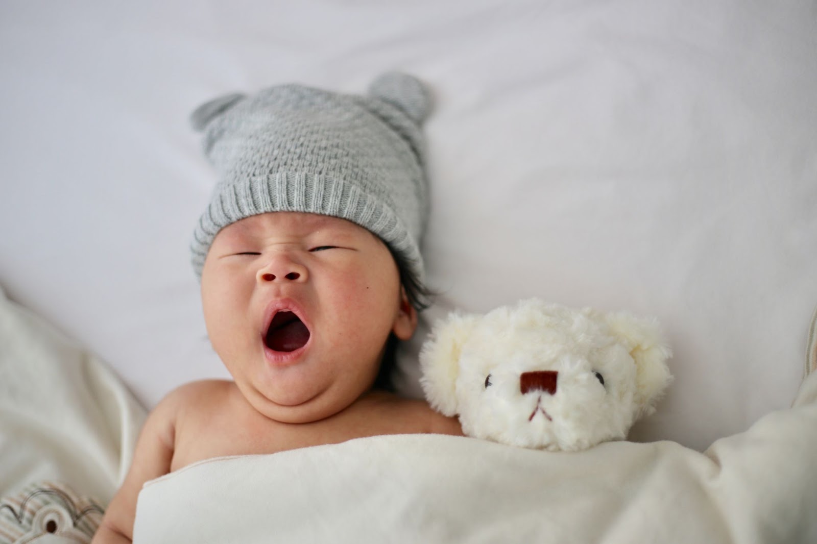An infant yawning in a bed with a teddy bear