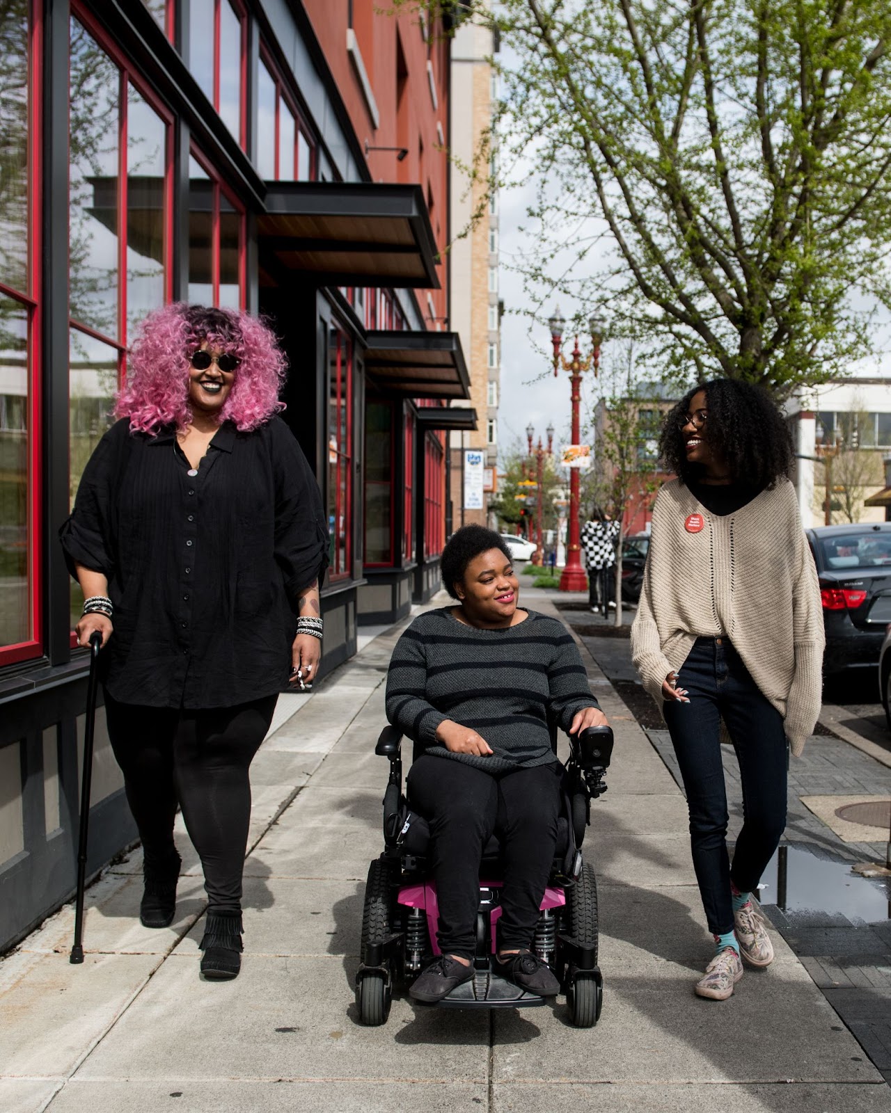 Three people on a sidewalk smiling at one another; one uses a cane, and one is riding in a wheelchair