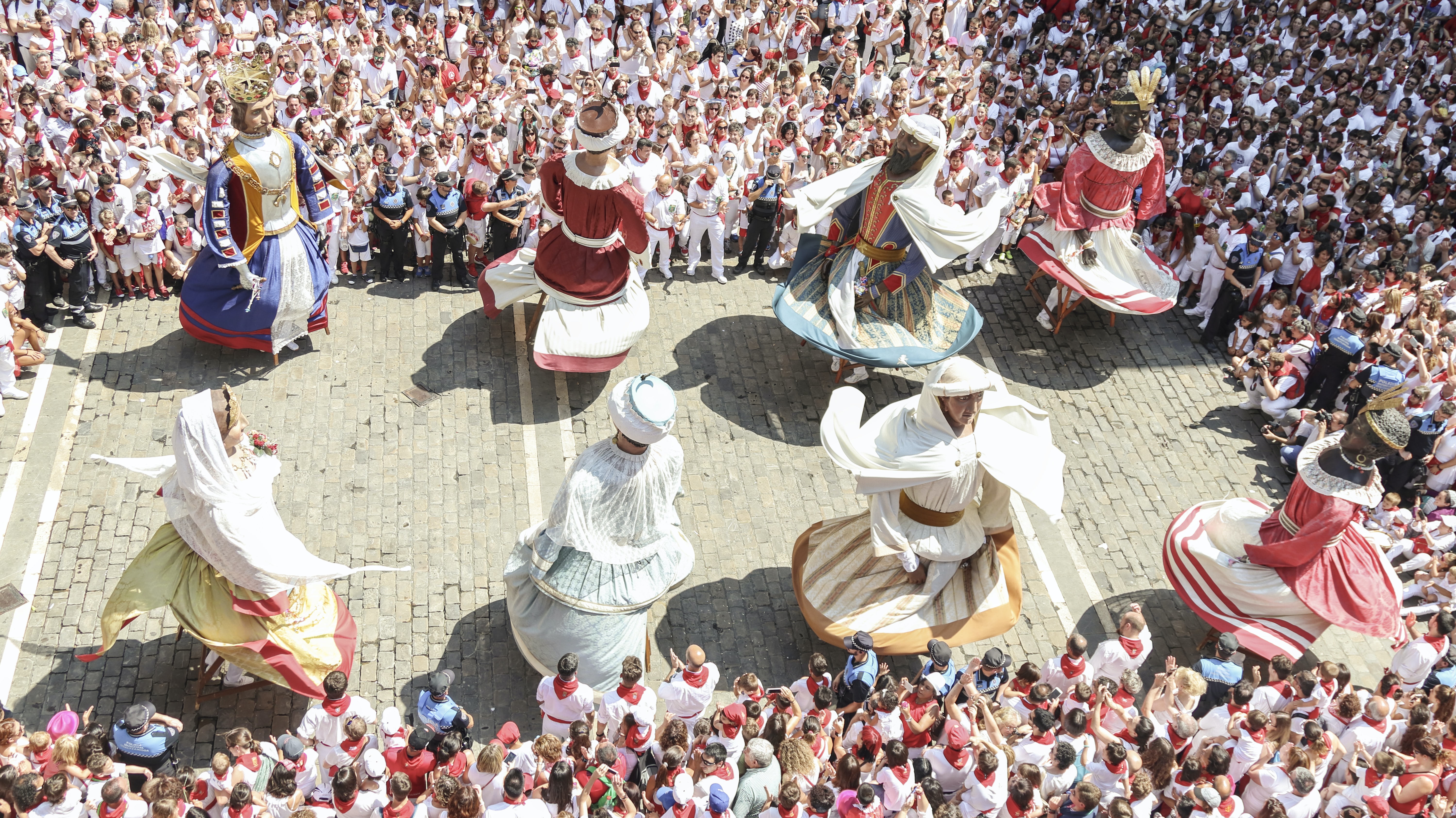 Aerial view of eight puppets with colorful dresses with white and brown hats in a town square, worn by people dancing