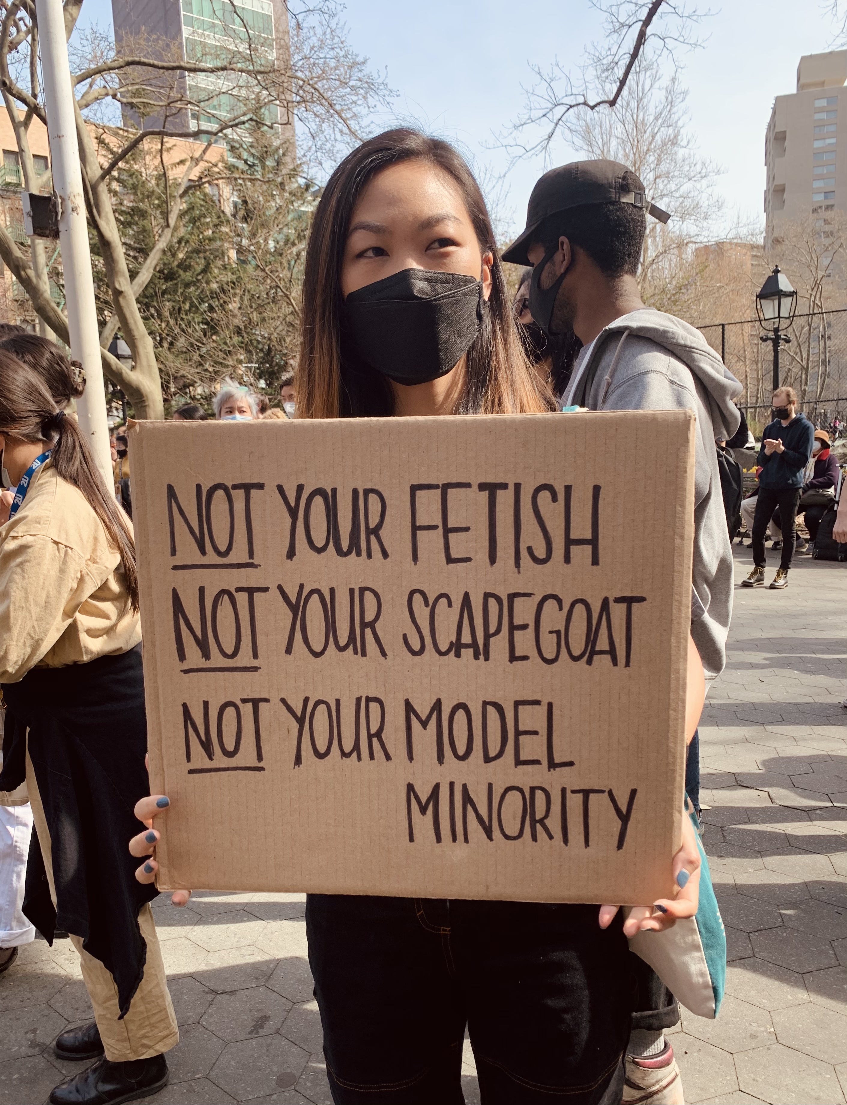 A masked protestor carrying a sign reading: Not your fetish, not your scapegoat, not your model minority