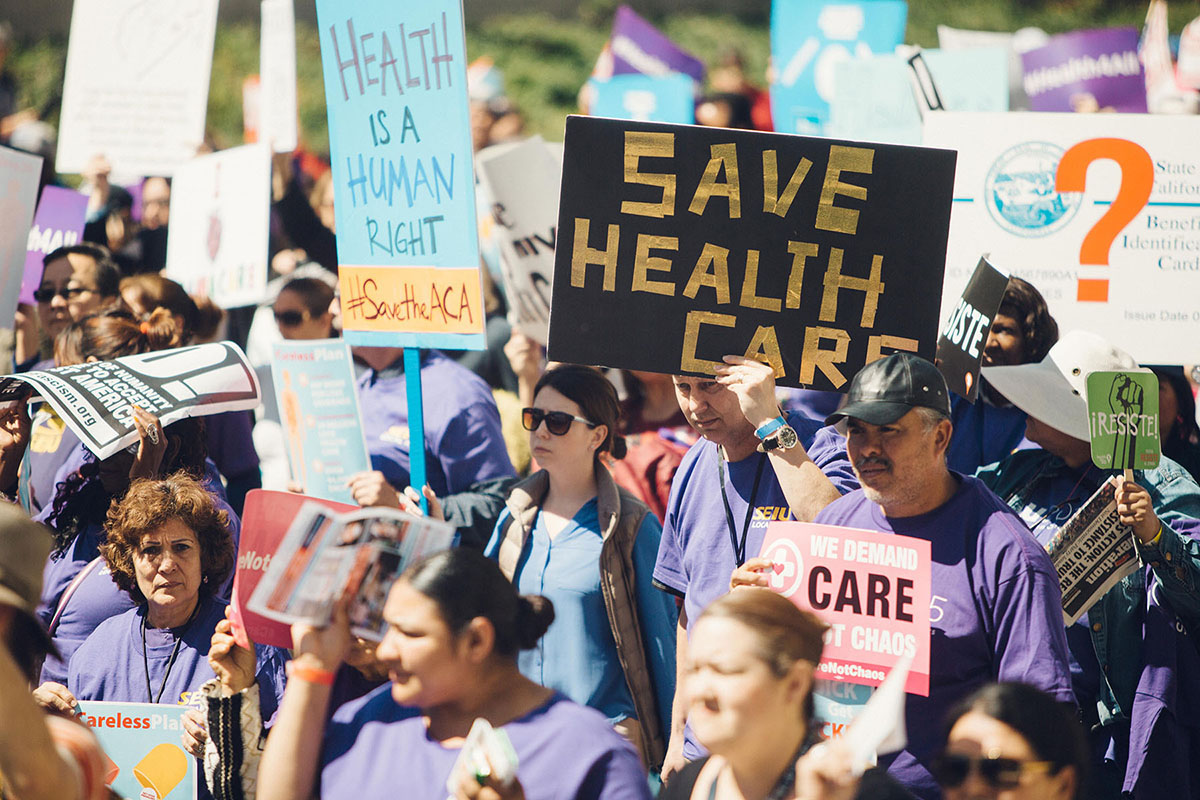 A large group of people gather at a rally. They hold signs reading Health is a Human Right and Save Health Care.