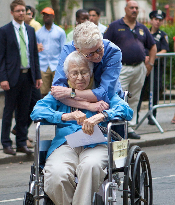 Phyllis Siegel and Connie Koplev embrace on a New York City street while people look on. Kopolev holds a document and sits in a wheelchair.