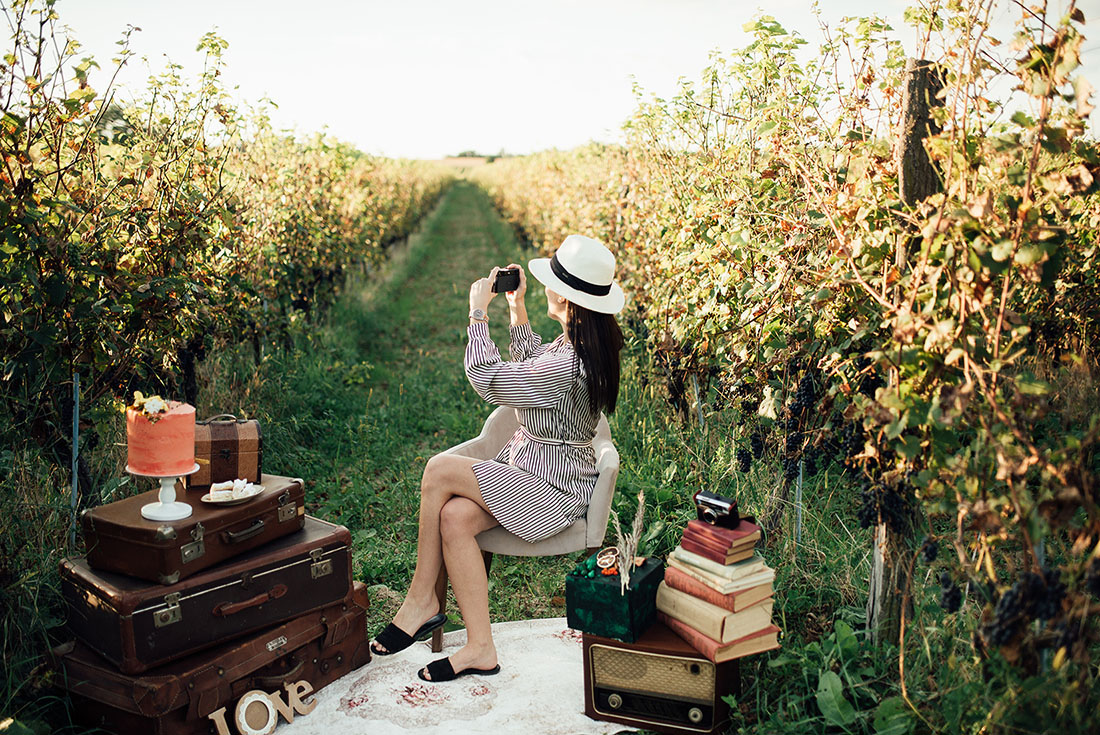 A person wearing a dress and with long hair sits in a vineyard with suitcases and old books set around them, as if for a photo shoot.