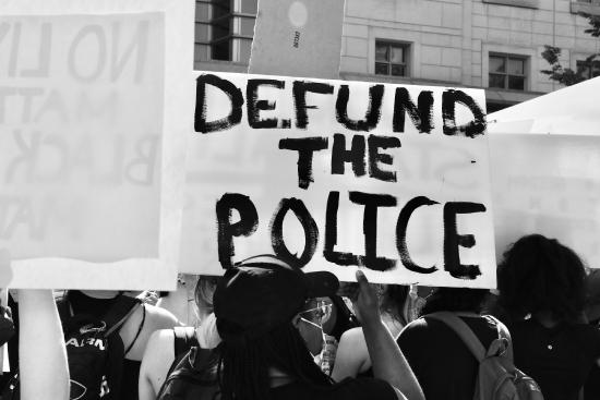 A black and white photo of the backs of protesters marching, one holding a sign Defund the Police