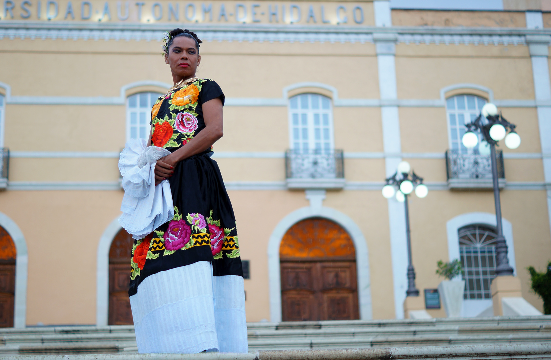 A Muxhe performer standing on steps, wearing a black and white dress with embroidered flowers in red, yellow, and purple