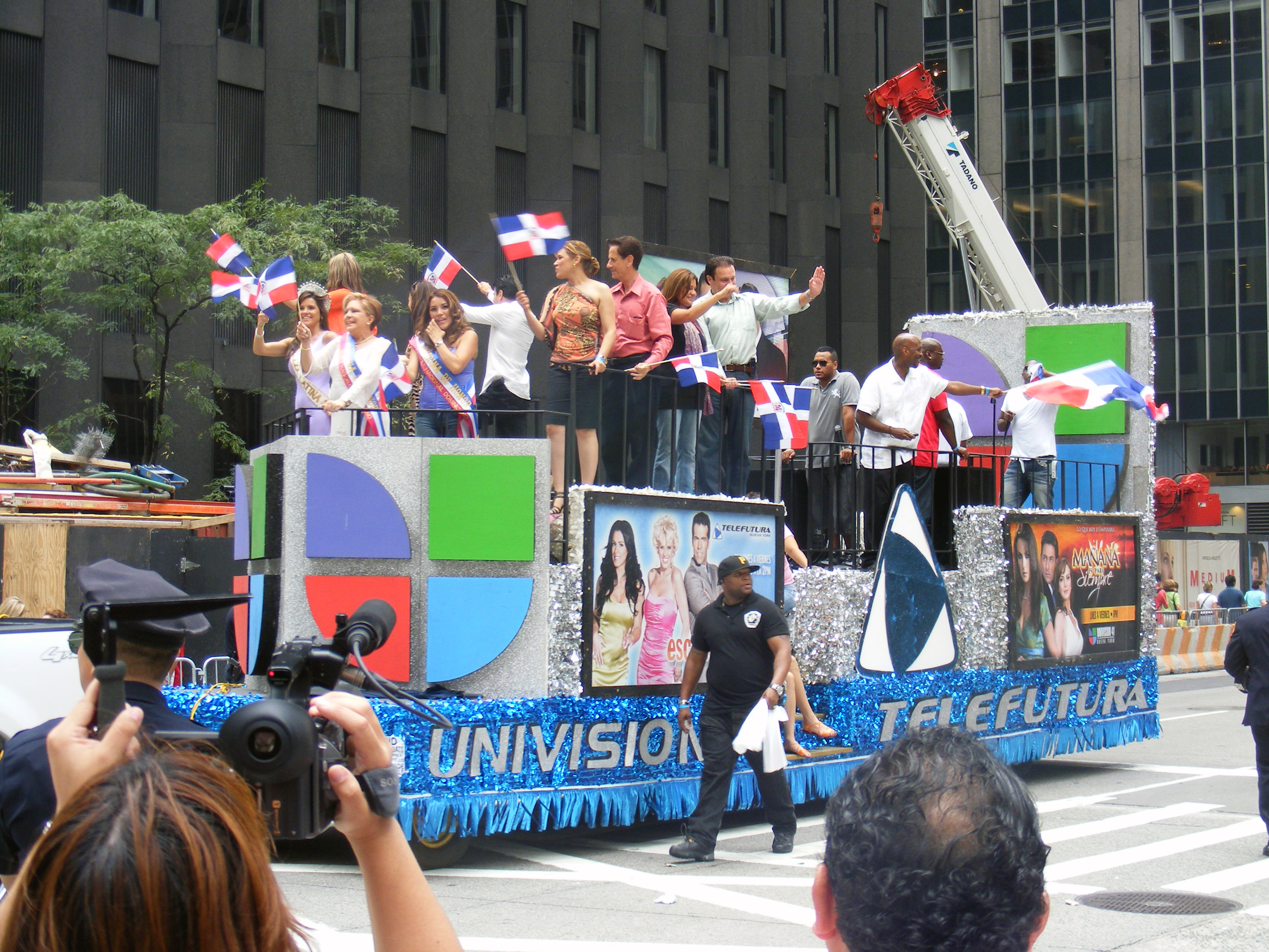 A group of adult people stand on a large float decorated with the Univision logo waving Dominican flags