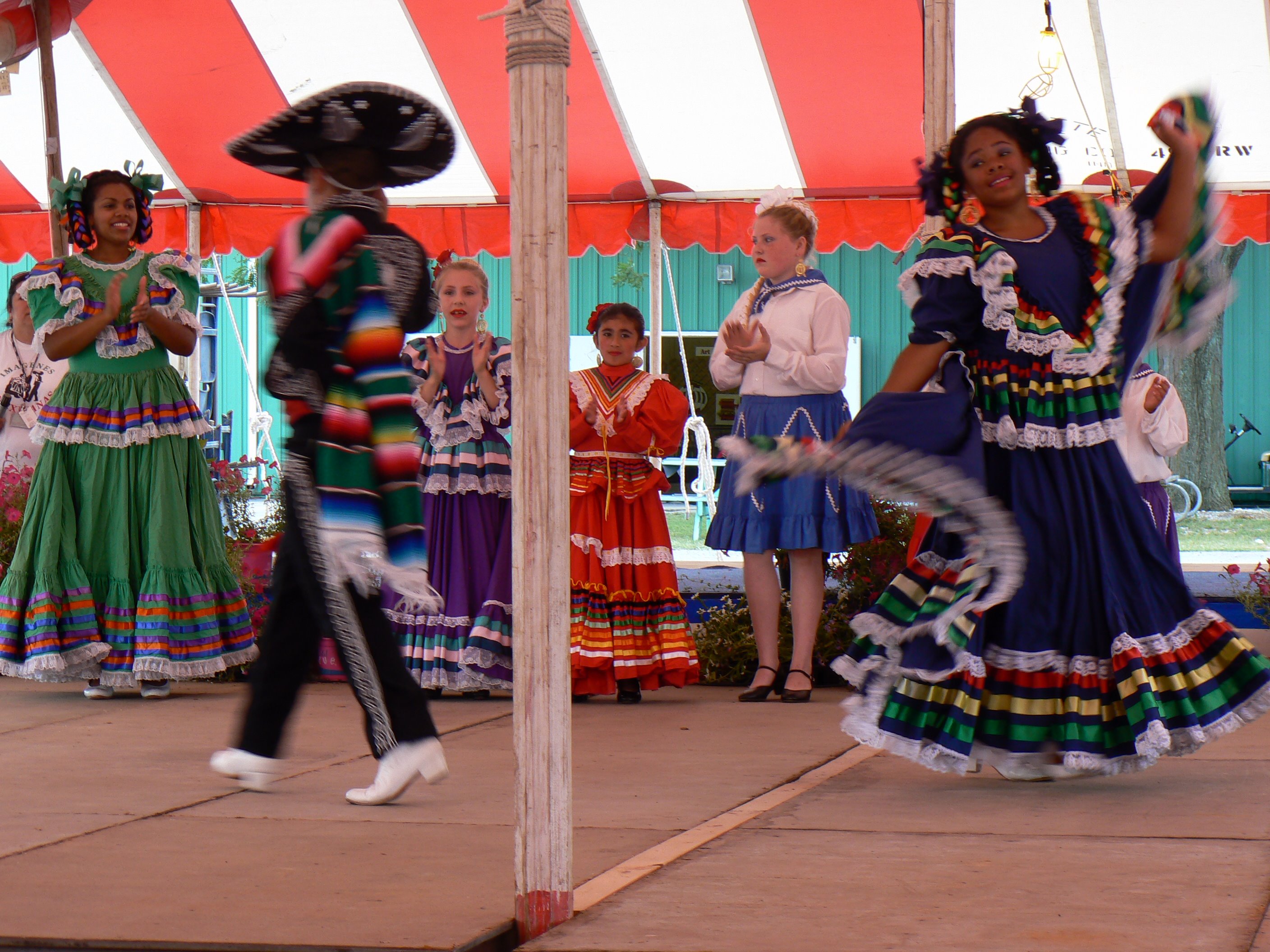 Two dancers performing ballet folklorico, with four others dressed in ballet folklorico dresses cheering them on.